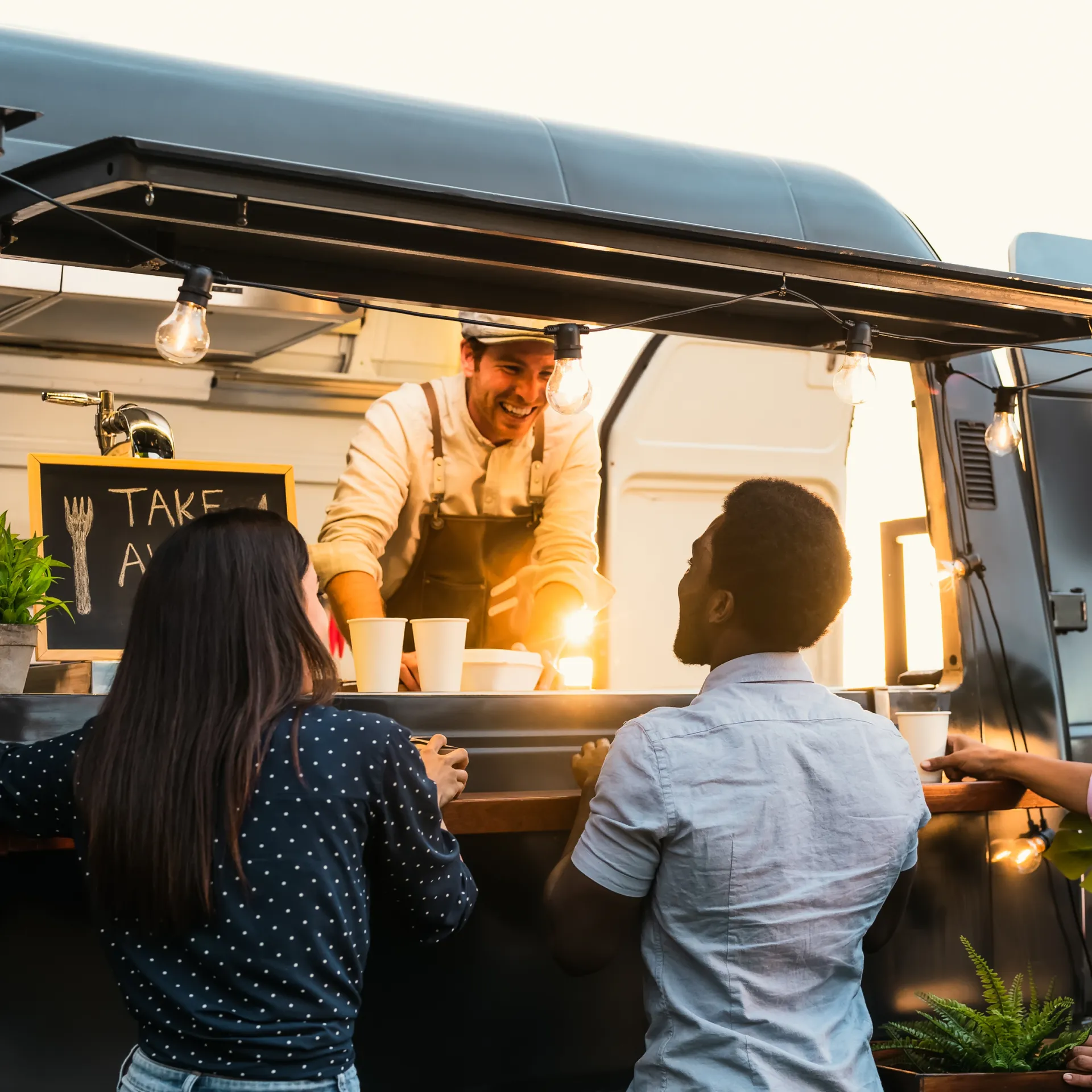 Three people stand at a food truck, engaging with a smiling vendor. Cups are on the counter under warm hanging lights.